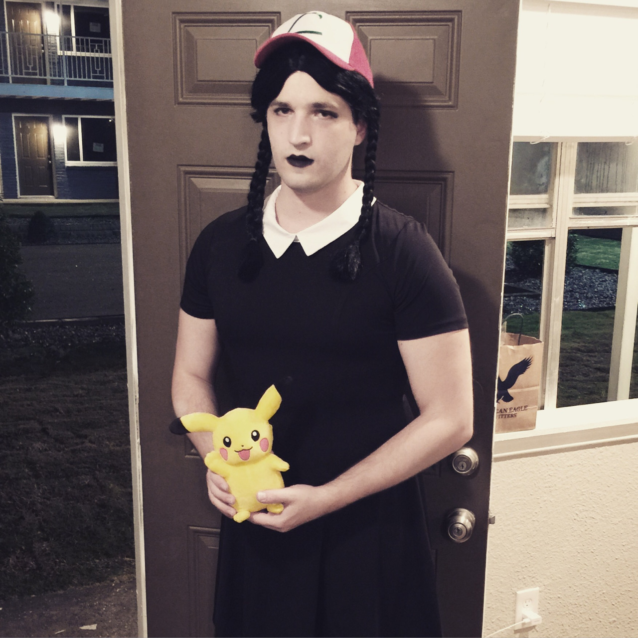A person cosplaying Wednesday Addams (black dress with white collar; black lips; pale face; dark long hairs braided in pigtails) wearing Ash Ketchum's cap holding a Pikachu doll.